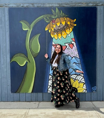 Artist stands in front of her sunflower-as-a-streetlight painting holding the light switch string that makes up the interactive element of the painting.
