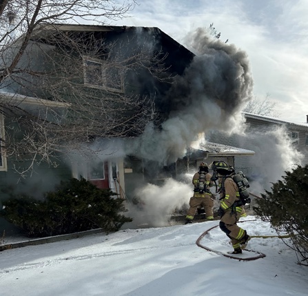 Smoke billows out of a burning house as two fire fighters hold the firehose and pump water.
